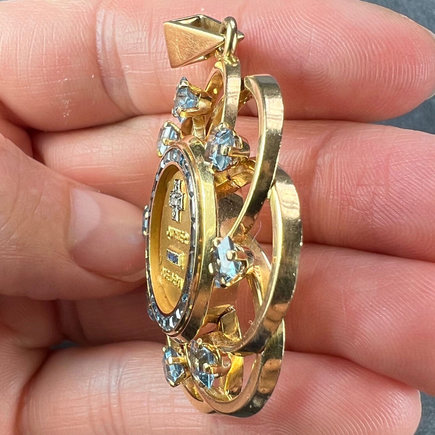 A yellow gold French QU'HIER QUE DEMAIN pendant with blue stones and diamonds, held in a hand on a grey background
