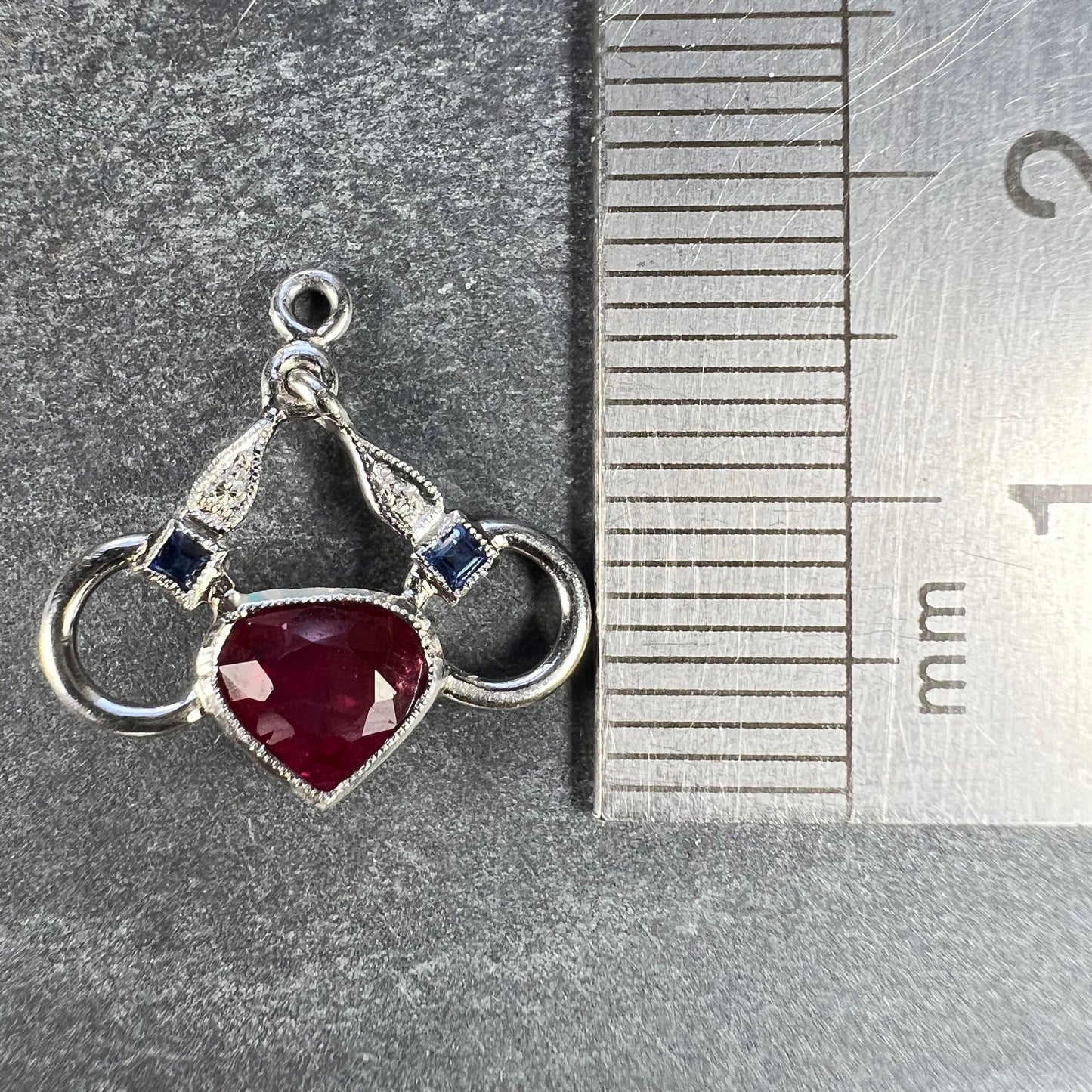 A heart shaped pendant with snaffle bits in white metal, with red, white and blue gemstones, next to a metal ruler on a grey background