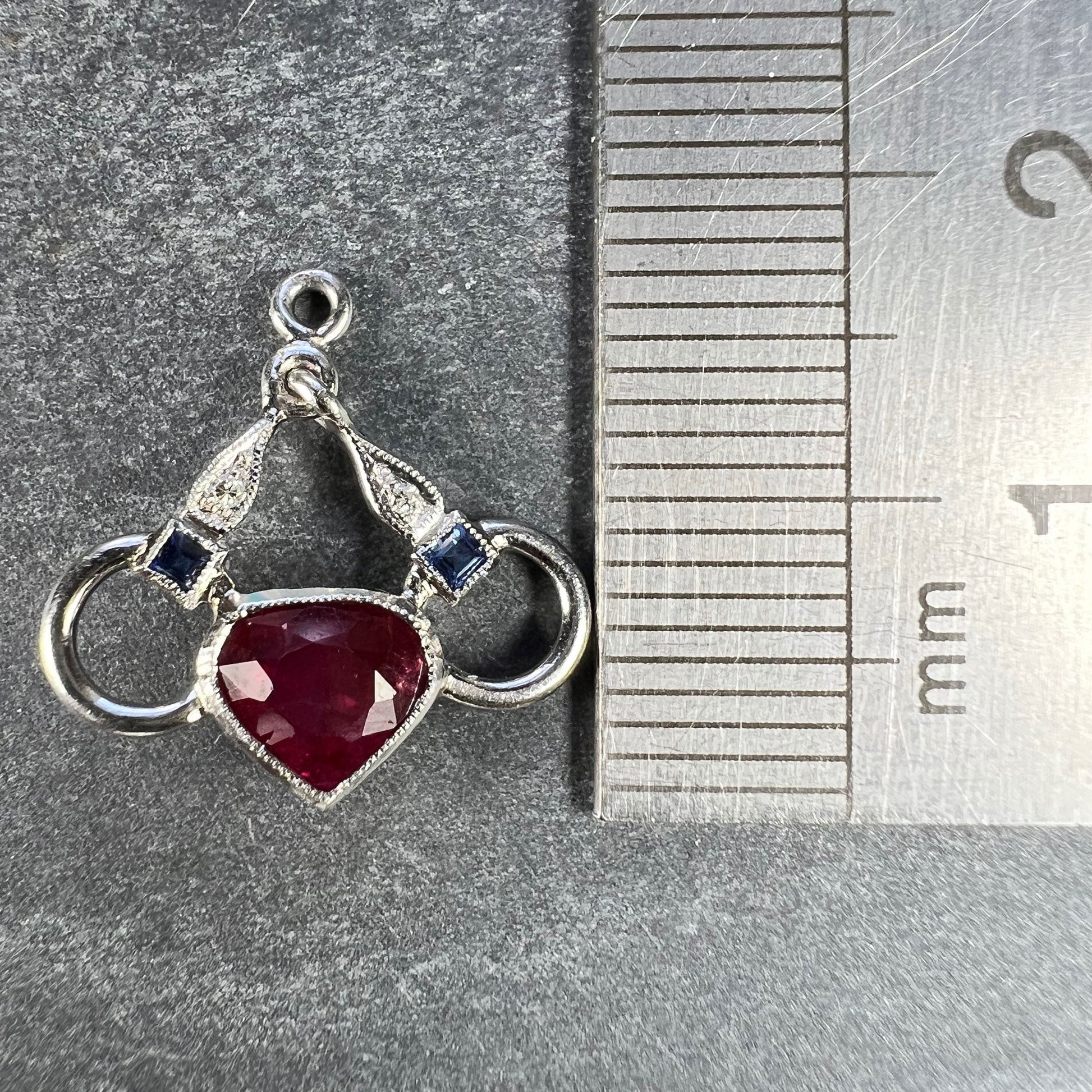 A heart shaped pendant with snaffle bits in white metal, with red, white and blue gemstones, next to a metal ruler on a grey background