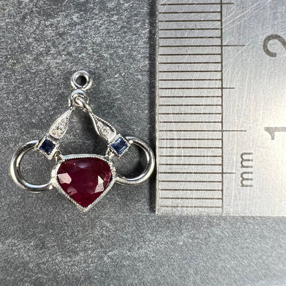 A heart shaped pendant with snaffle bits in white metal, with red, white and blue gemstones, next to a metal ruler on a grey background