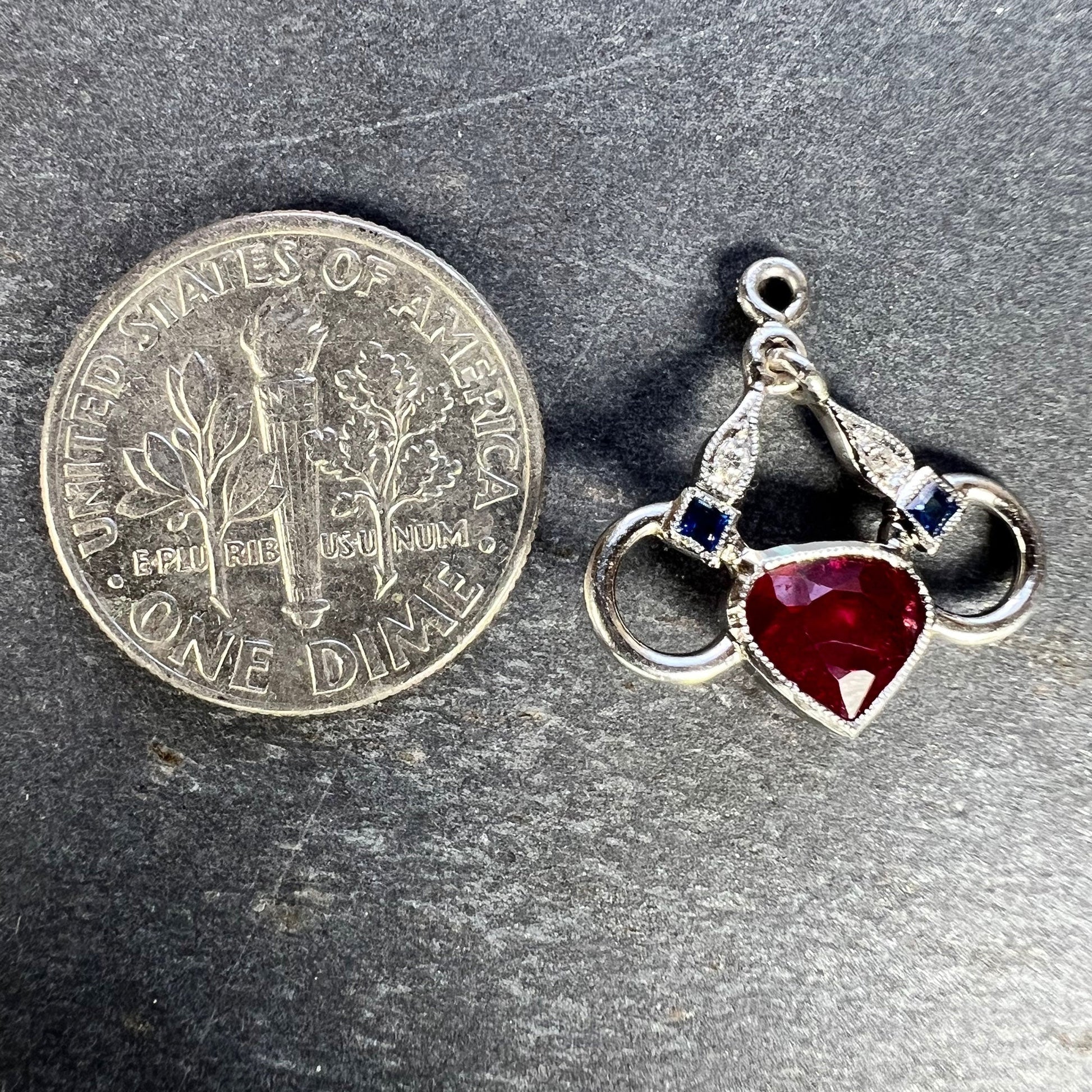 A heart shaped pendant with snaffle bits in white metal, with red, white and blue gemstones, next to a dime coin on a grey background