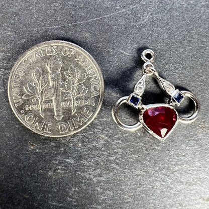 A heart shaped pendant with snaffle bits in white metal, with red, white and blue gemstones, next to a dime coin on a grey background