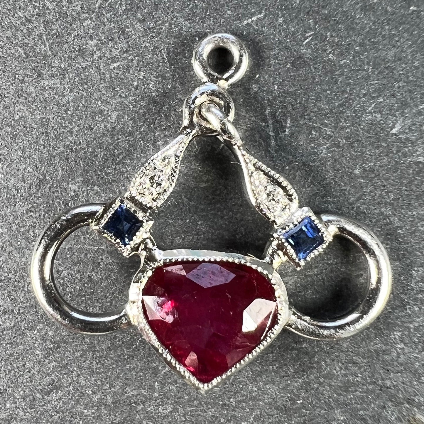 A heart shaped pendant with snaffle bits in white metal, with red, white and blue gemstones, on a grey background