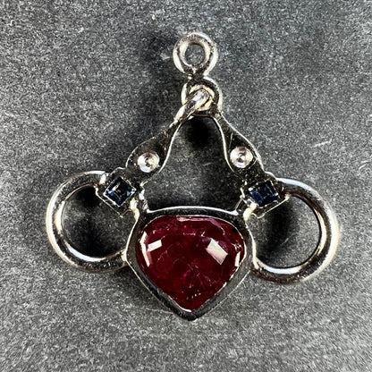 A heart shaped pendant with snaffle bits in white metal, with red, white and blue gemstones, on a grey background