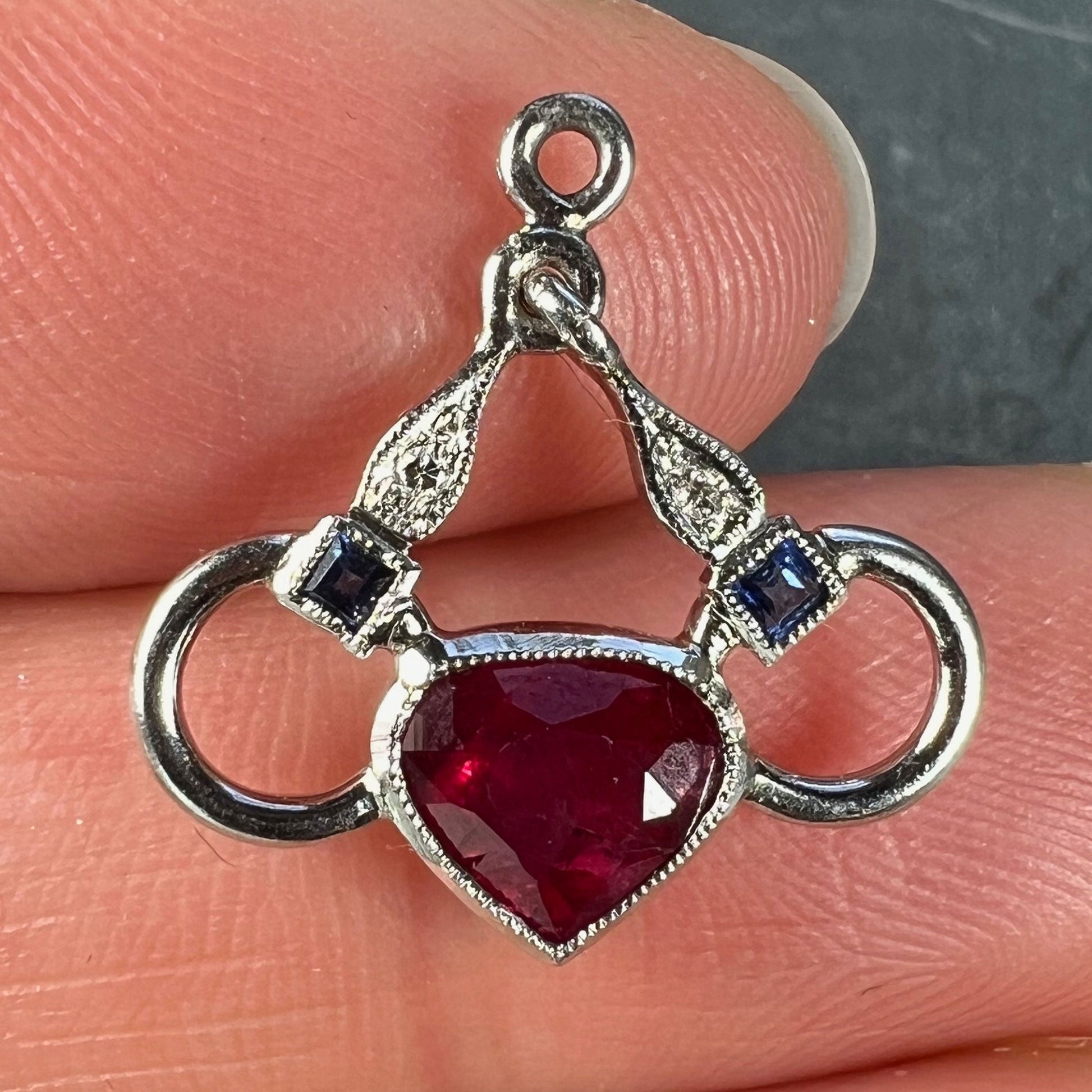 A heart shaped pendant with snaffle bits in white metal, with red, white and blue gemstones, held in a hand on a grey background