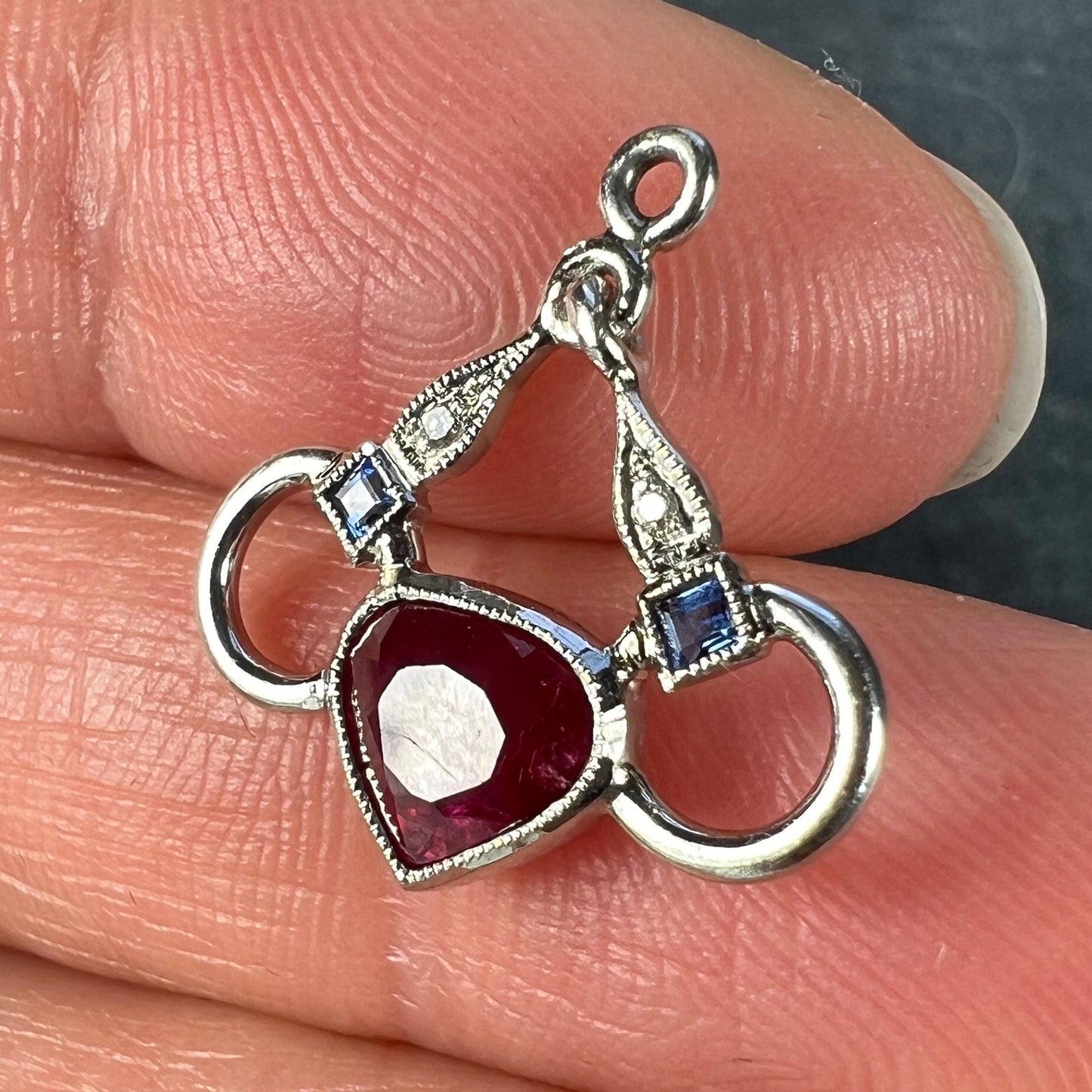 A heart shaped pendant with snaffle bits in white metal, with red, white and blue gemstones, held in a hand on a grey background