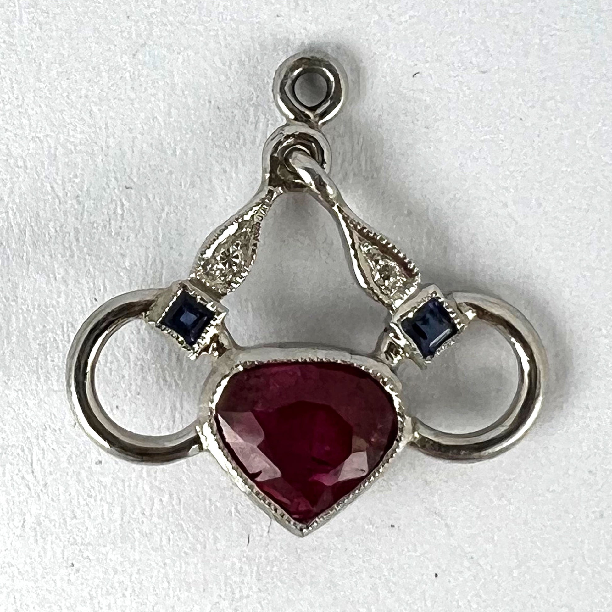 A heart shaped pendant with snaffle bits in white metal, with red, white and blue gemstones, on a white background