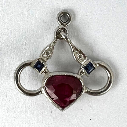 A heart shaped pendant with snaffle bits in white metal, with red, white and blue gemstones, on a white background