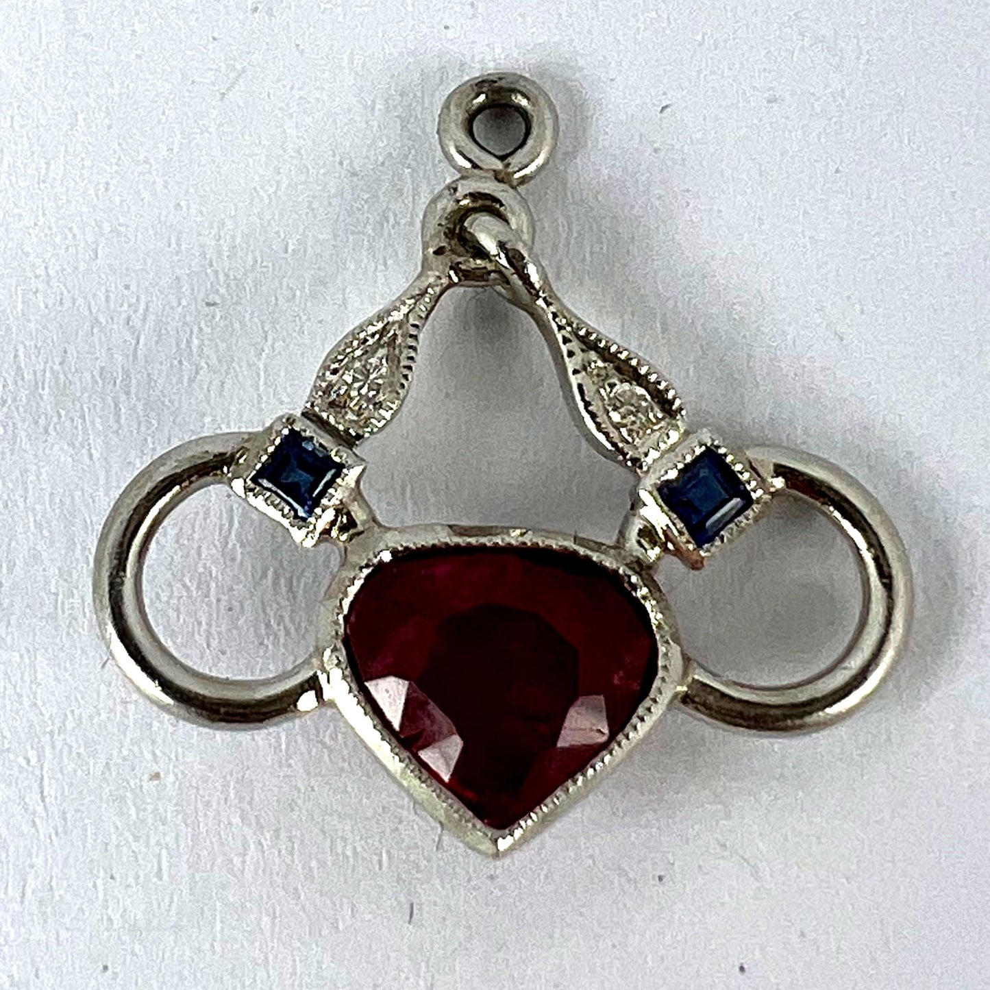 A heart shaped pendant with snaffle bits in white metal, with red, white and blue gemstones, on a white background