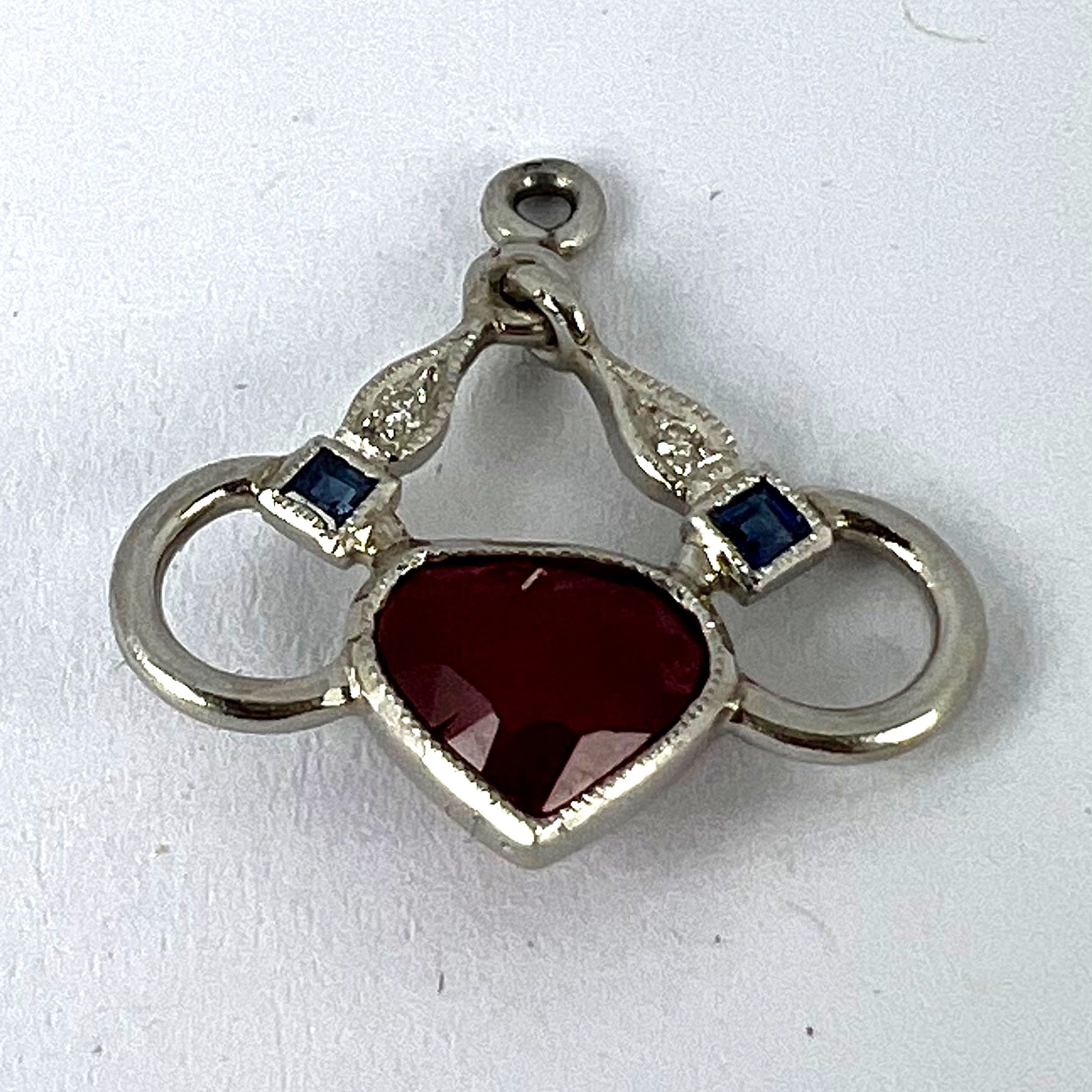 A heart shaped pendant with snaffle bits in white metal, with red, white and blue gemstones, on a white background