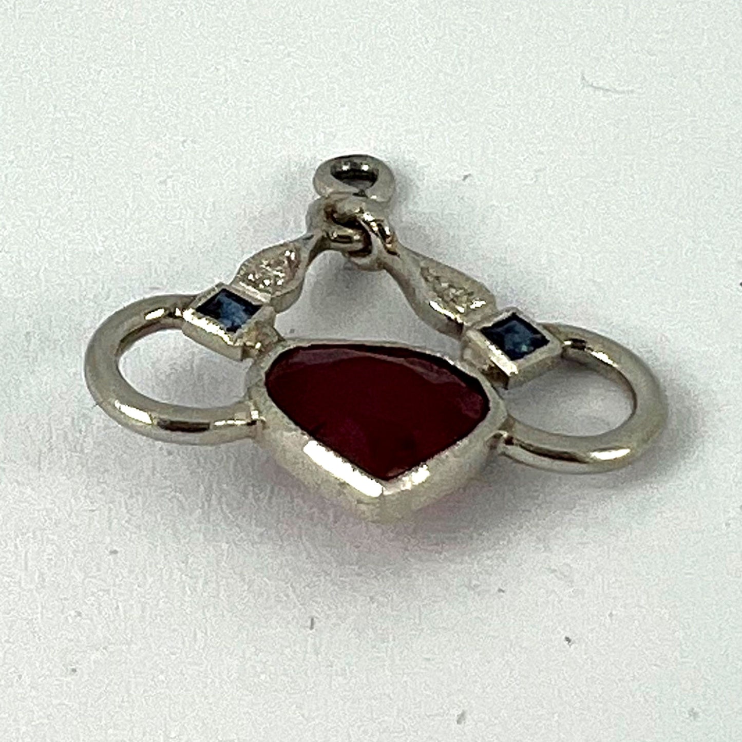 A heart shaped pendant with snaffle bits in white metal, with red, white and blue gemstones, on a white background