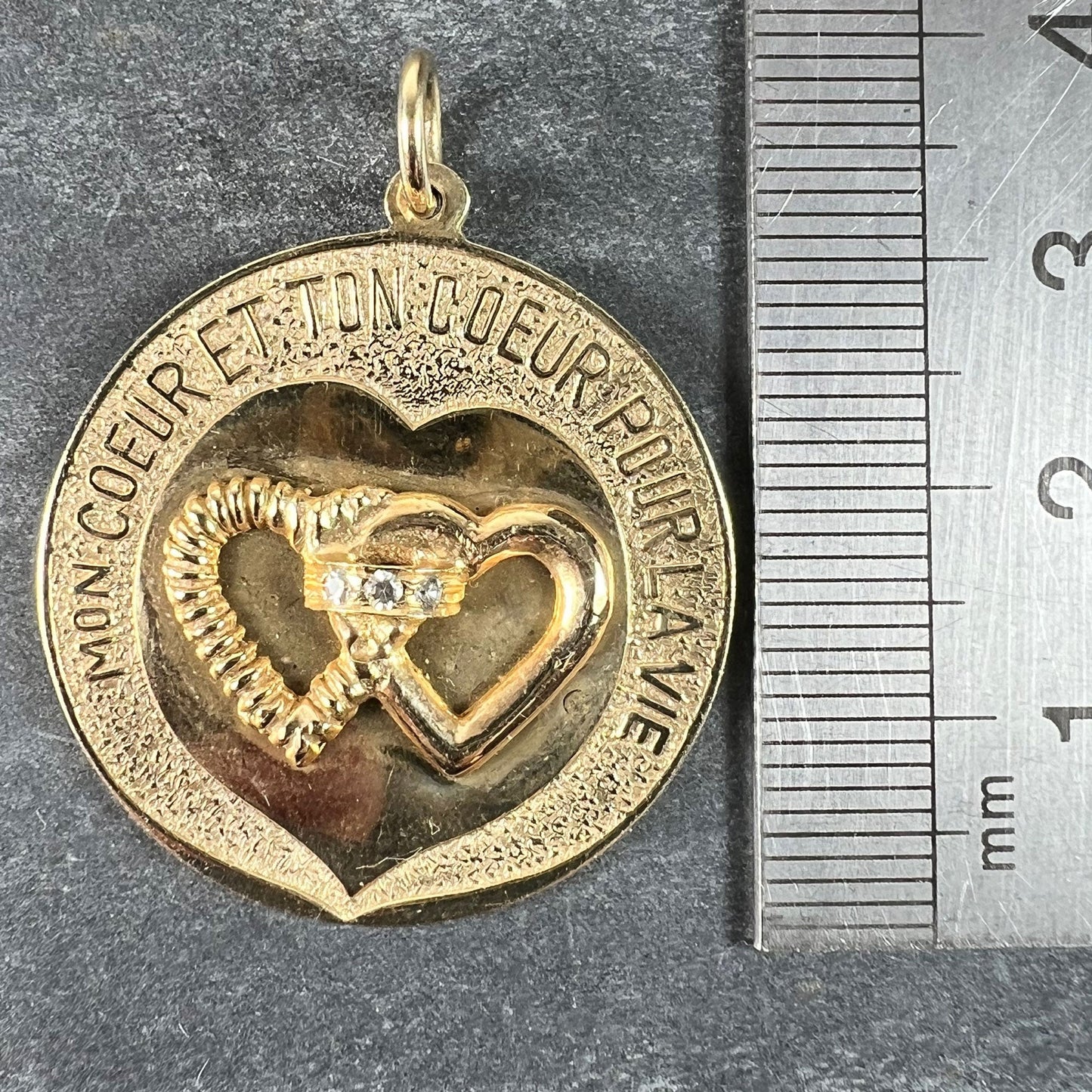 A round yellow gold love heart diamond pendant engraved MON COEUR ET TON COEUR POUR LA VIE, next to a metal ruler
