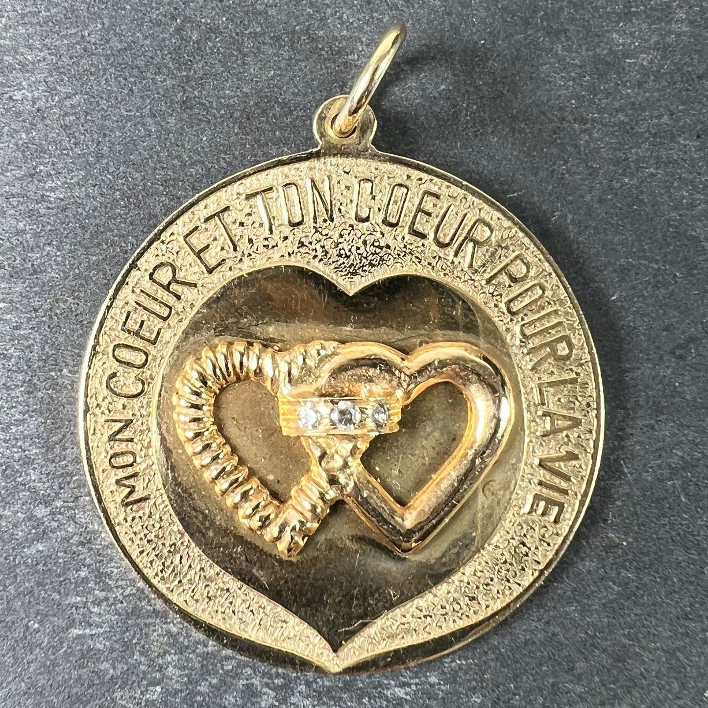 A round yellow gold love heart diamond pendant engraved MON COEUR ET TON COEUR POUR LA VIE on a grey background