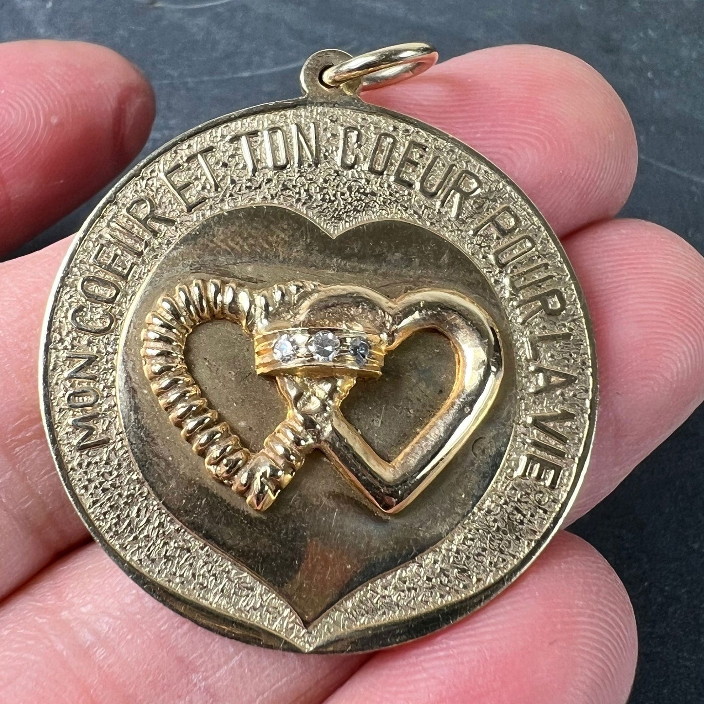 A round yellow gold love heart diamond pendant engraved MON COEUR ET TON COEUR POUR LA VIE, held in a hand