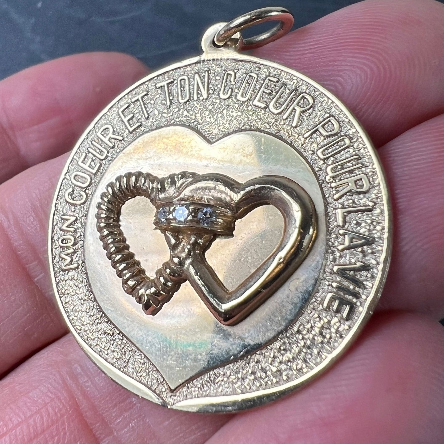 A round yellow gold love heart diamond pendant engraved MON COEUR ET TON COEUR POUR LA VIE, held in a hand