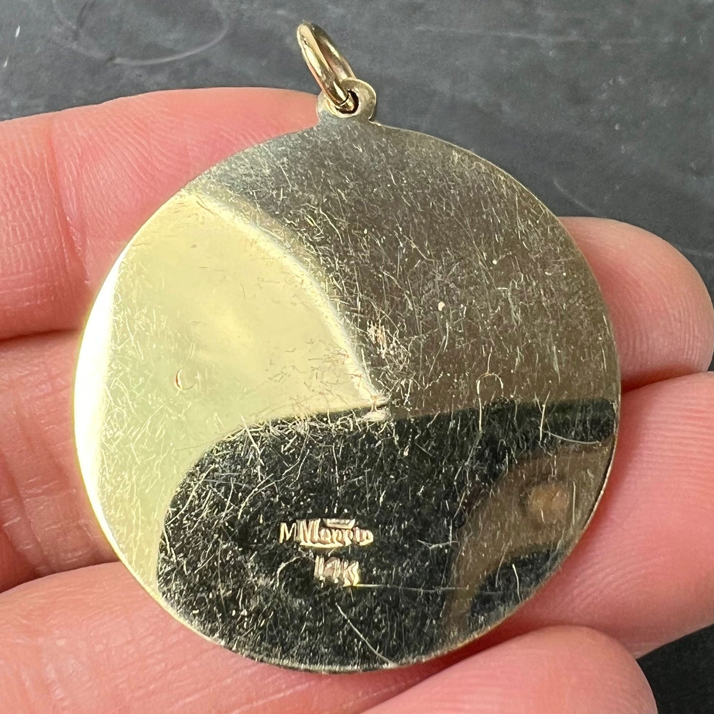 A round yellow gold pendant stamped Merrin and 14K, held in a hand