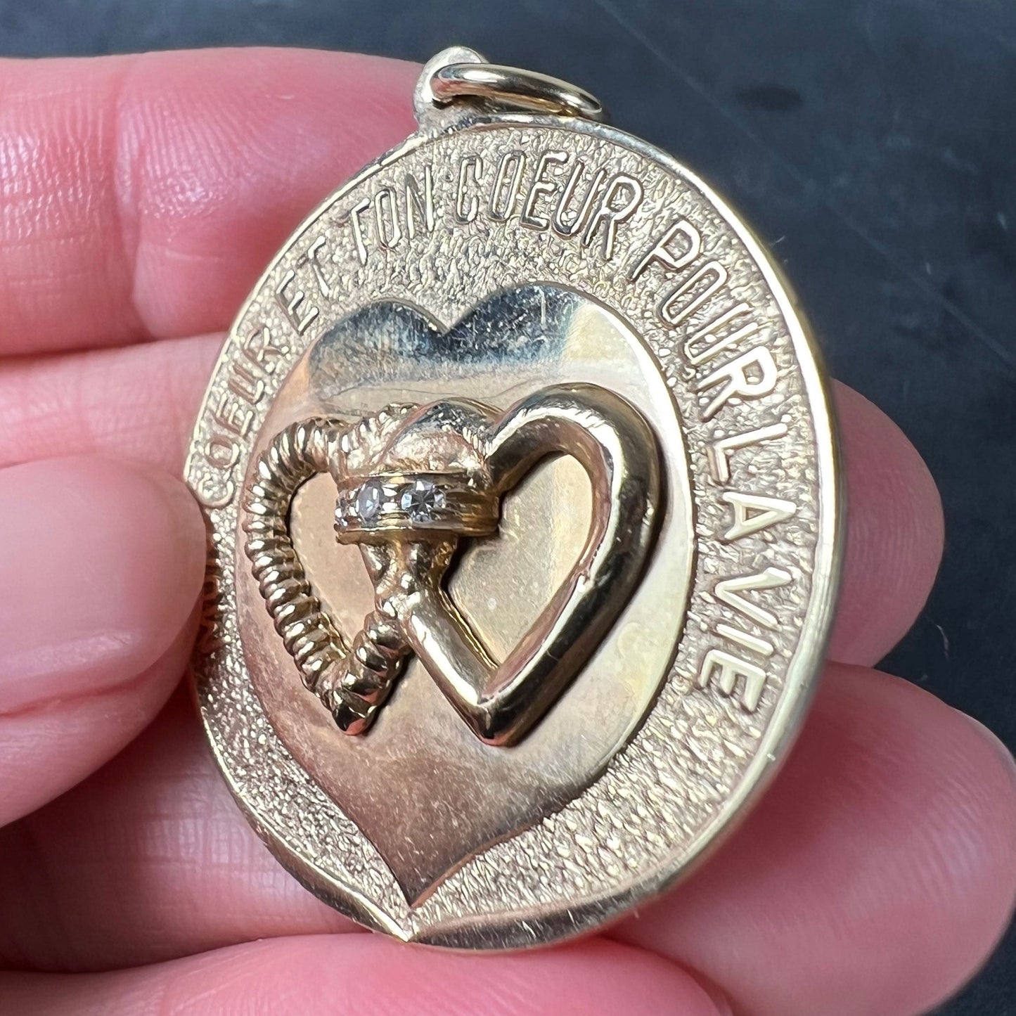 A round yellow gold love heart diamond pendant engraved MON COEUR ET TON COEUR POUR LA VIE, held in a hand