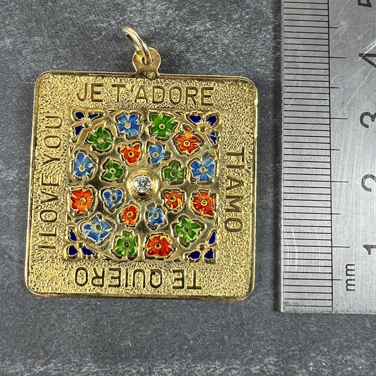 A square shaped yellow gold pendant with enamelled flowers and a central diamond, with text reading JE T'ADORE, TI AMO, TE QUIERO, I LOVE YOU, next to a metal ruler