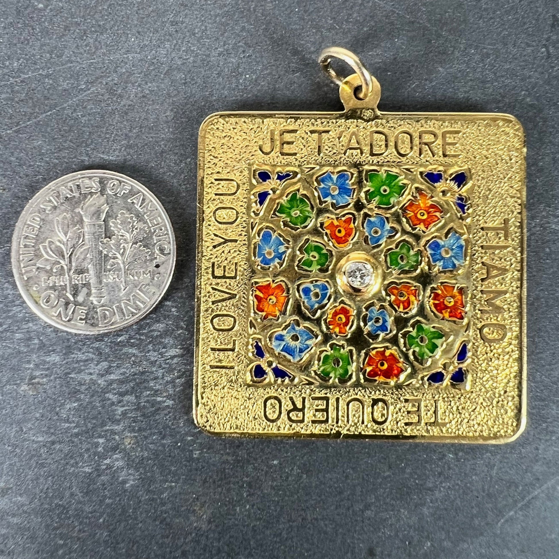 A square shaped yellow gold pendant with enamelled flowers and a central diamond, with text reading JE T'ADORE, TI AMO, TE QUIERO, I LOVE YOU, next to a dime coin