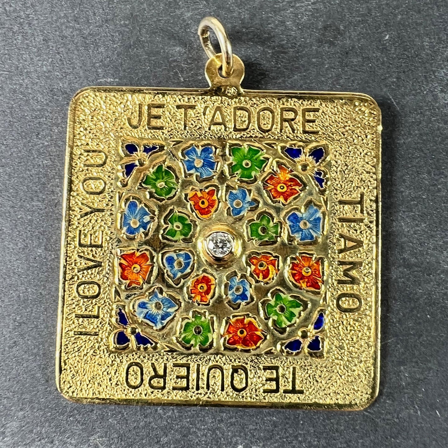 A square shaped yellow gold pendant with enamelled flowers and a central diamond, with text reading JE T'ADORE, TI AMO, TE QUIERO, I LOVE YOU