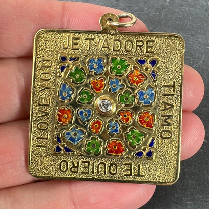 A square shaped yellow gold pendant with enamelled flowers and a central diamond, with text reading JE T'ADORE, TI AMO, TE QUIERO, I LOVE YOU, held in a hand
