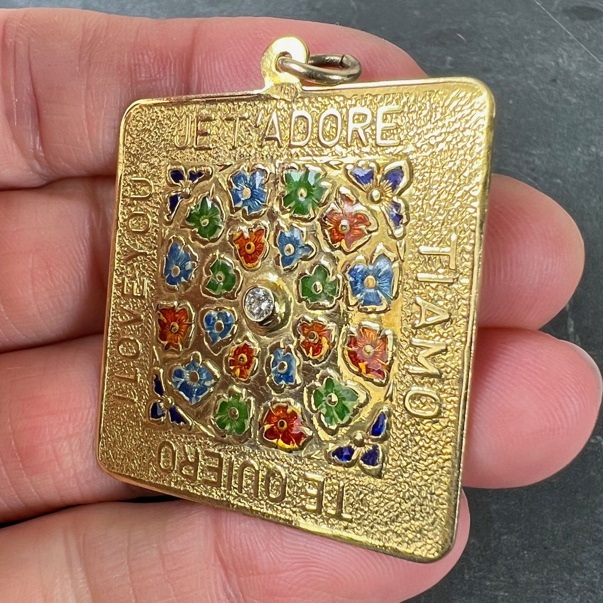 A square shaped yellow gold pendant with enamelled flowers and a central diamond, with text reading JE T'ADORE, TI AMO, TE QUIERO, I LOVE YOU, held in a hand