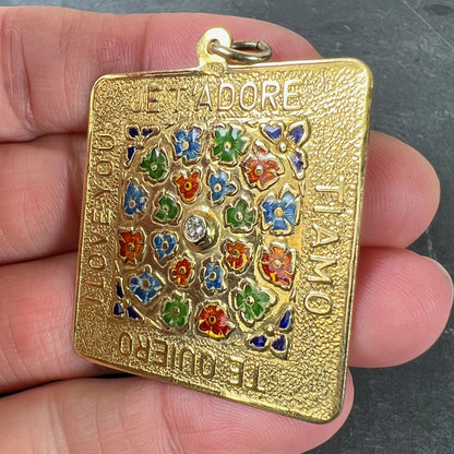 A square shaped yellow gold pendant with enamelled flowers and a central diamond, with text reading JE T'ADORE, TI AMO, TE QUIERO, I LOVE YOU, held in a hand