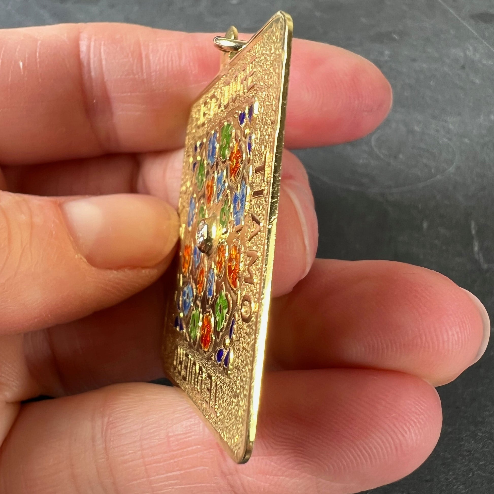 A square shaped yellow gold pendant with enamelled flowers and a central diamond, with text reading JE T'ADORE, TI AMO, TE QUIERO, I LOVE YOU, held in a hand