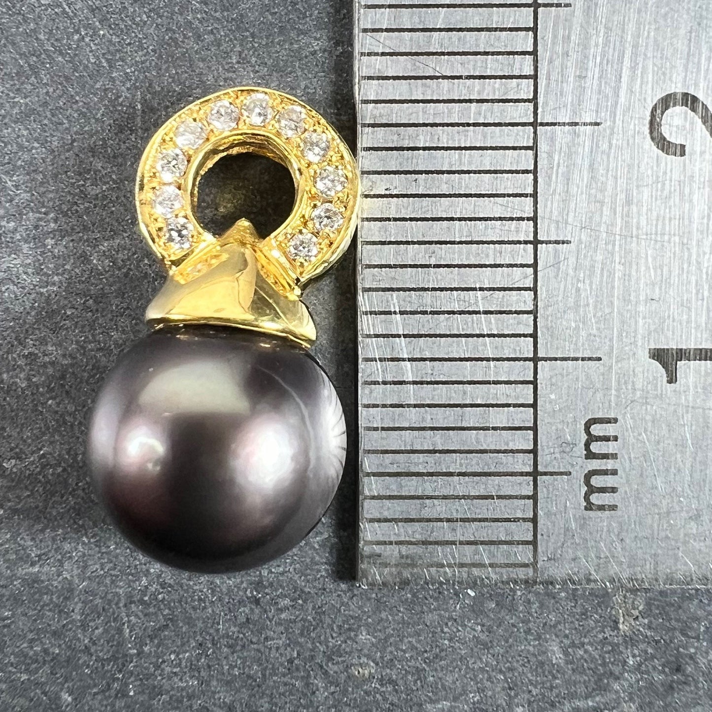 A black pearl pendant with a yellow gold bale set with diamonds, next to a metal ruler on a grey background