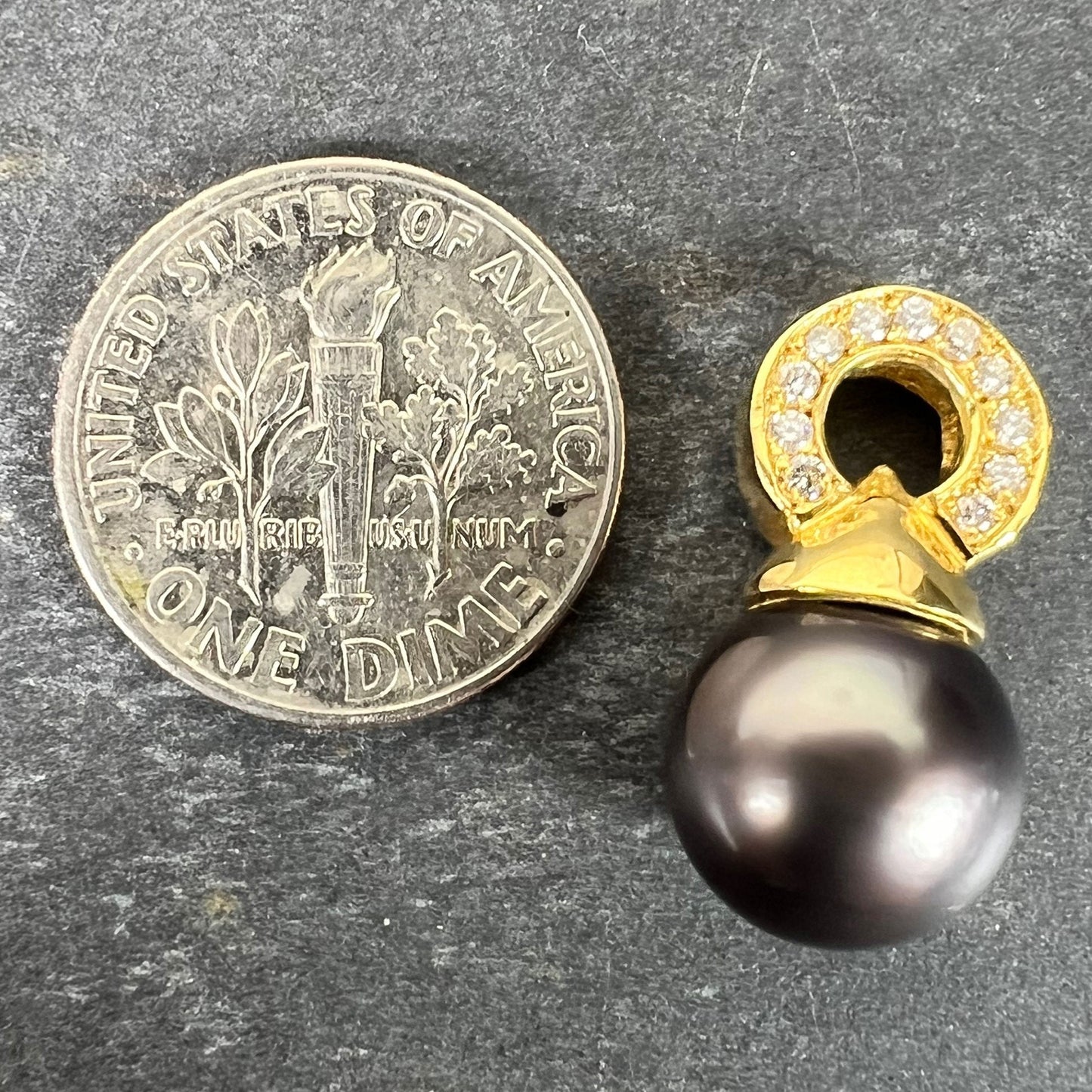 A black pearl pendant with a yellow gold bale set with diamonds, next to a dime coin on a grey background
