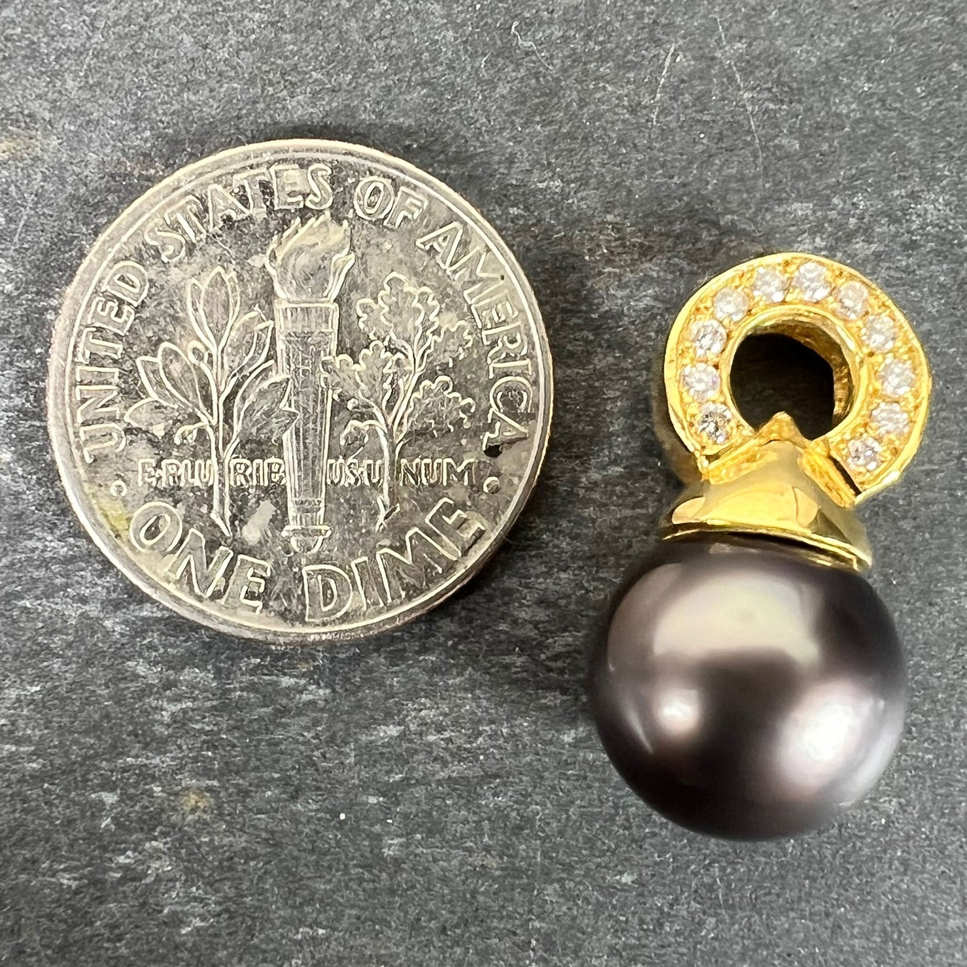 A black pearl pendant with a yellow gold bale set with diamonds, next to a dime coin on a grey background