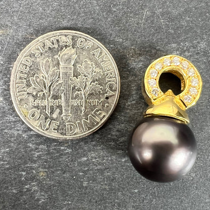 A black pearl pendant with a yellow gold bale set with diamonds, next to a dime coin on a grey background