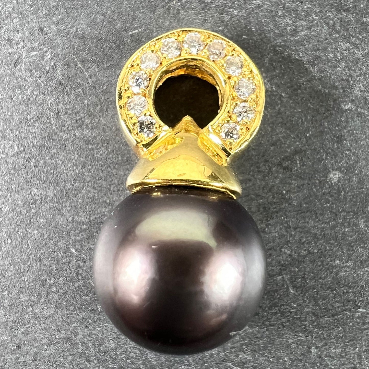 A black pearl pendant with a yellow gold bale set with diamonds, on a grey background
