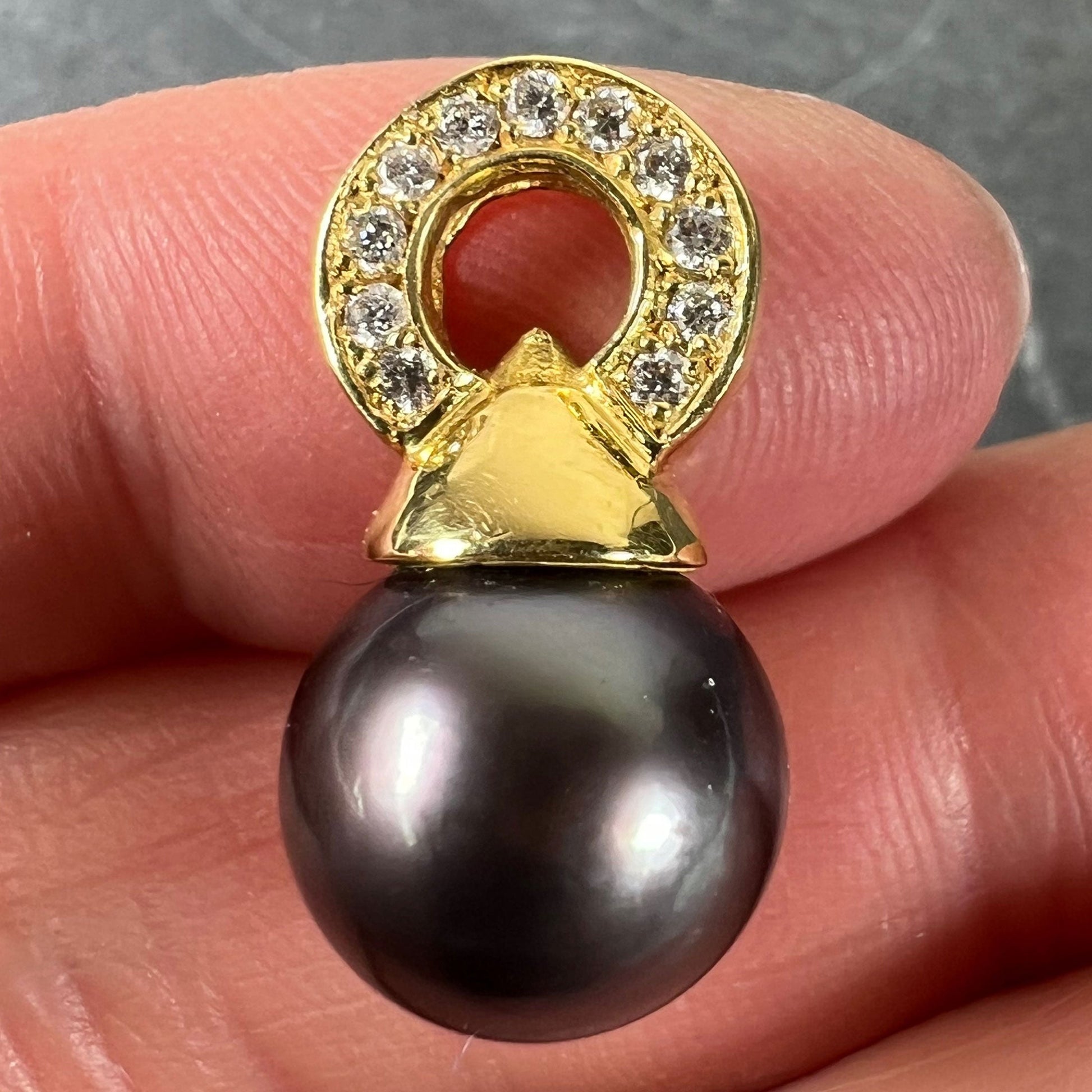 A black pearl pendant with a yellow gold bale set with diamonds, held in a hand on a grey background