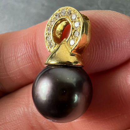 A black pearl pendant with a yellow gold bale set with diamonds, held in a hand on a grey background