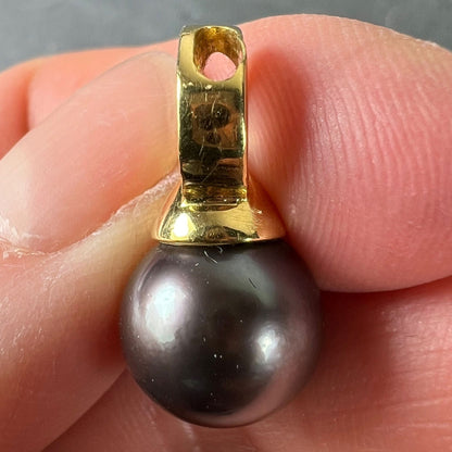 A black pearl pendant with a yellow gold bale set with diamonds, held in a hand on a grey background