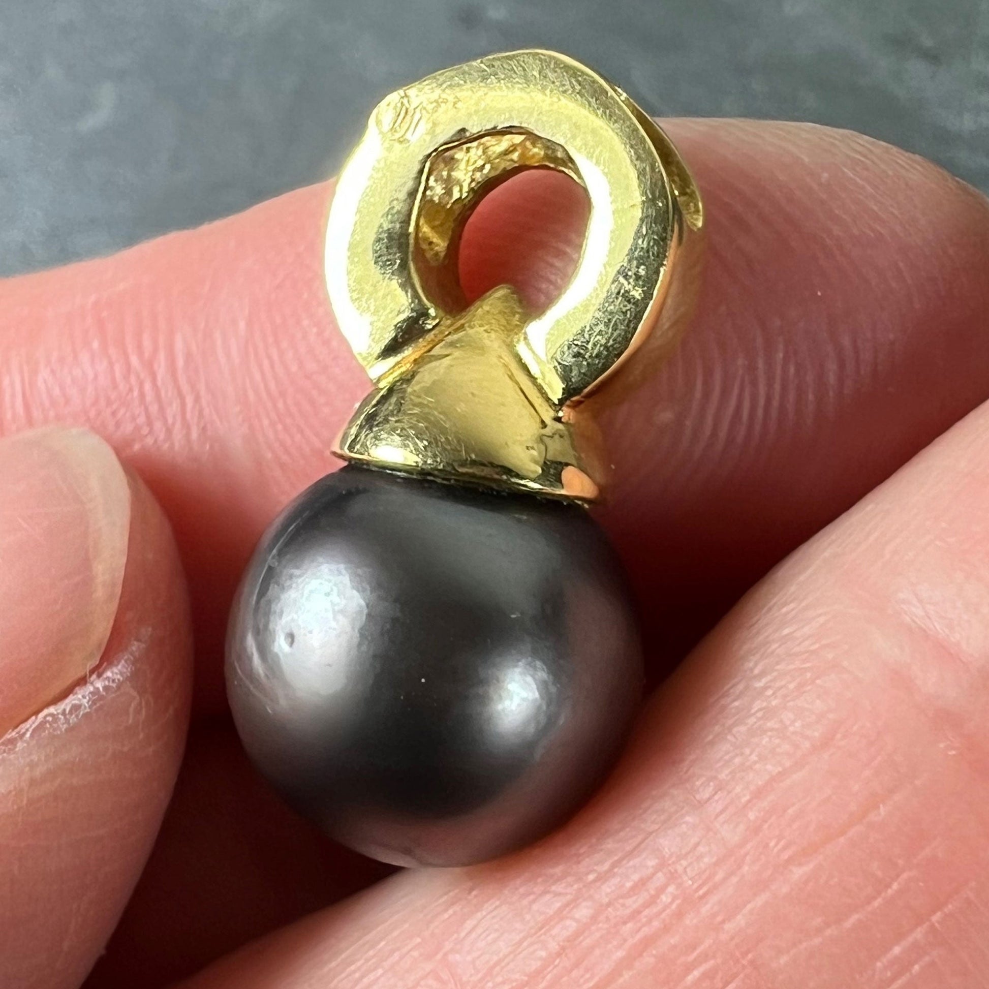 A black pearl pendant with a yellow gold bale, held in a hand on a grey background