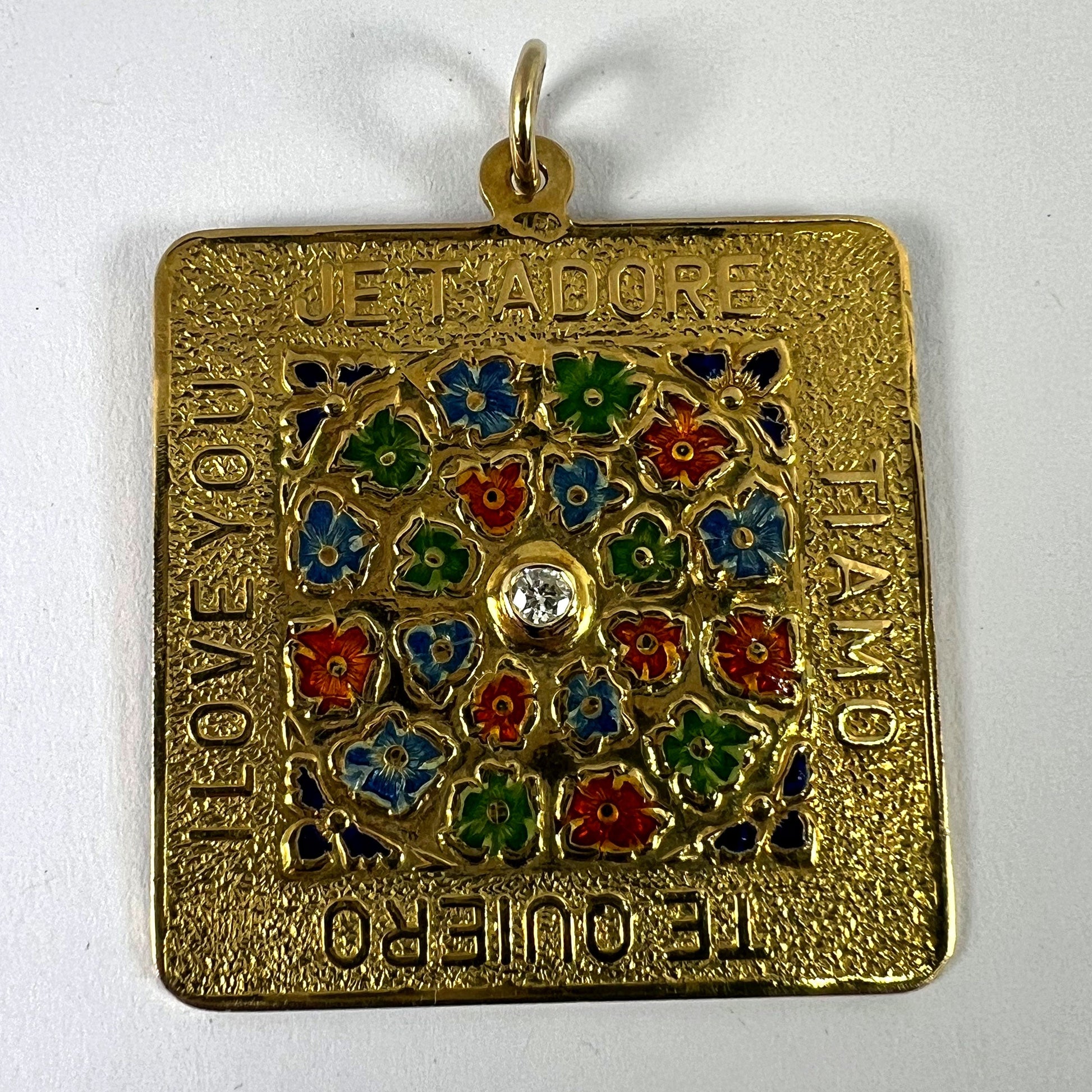 A square shaped yellow gold pendant with enamelled flowers and a central diamond, with text reading JE T'ADORE, TI AMO, TE QUIERO, I LOVE YOU