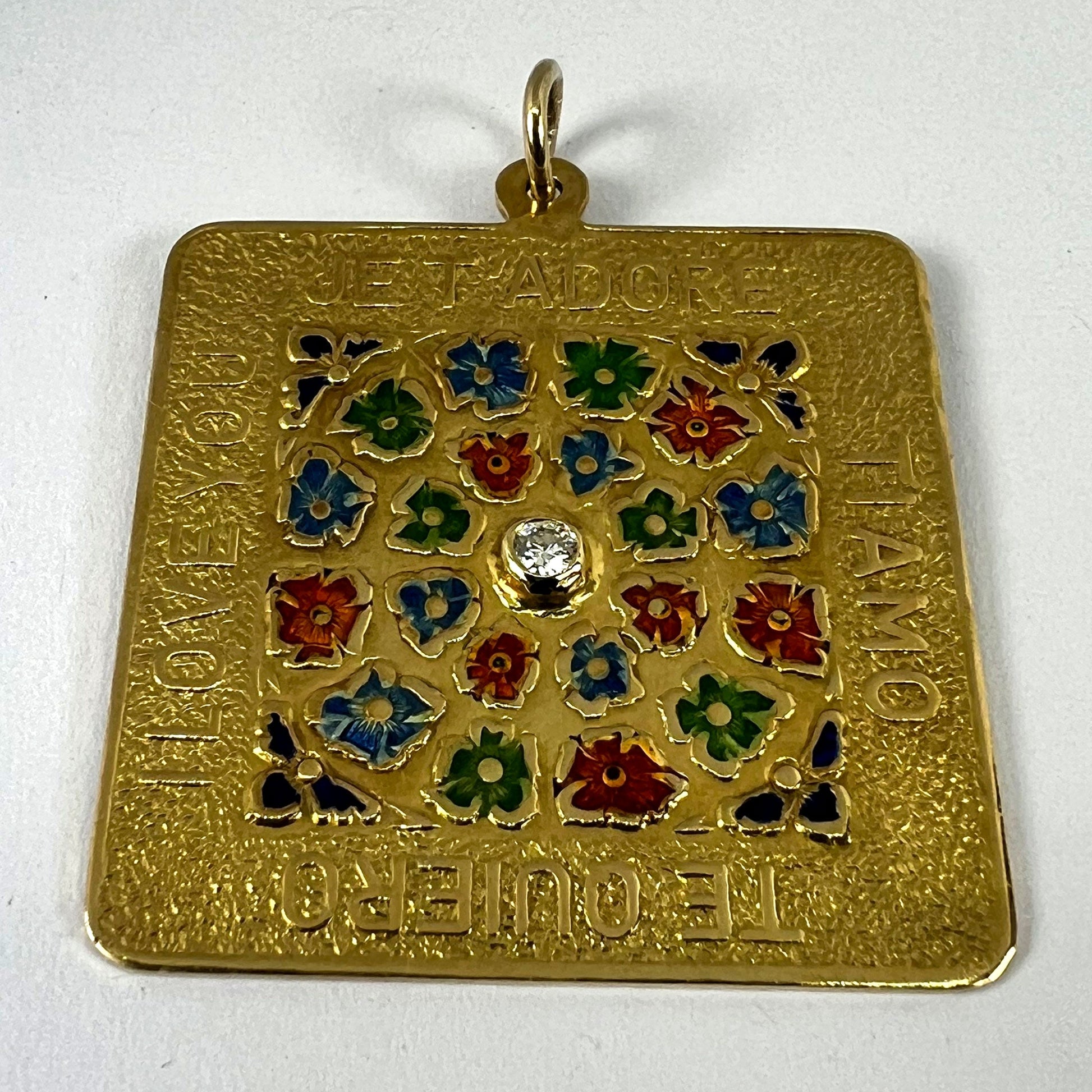 A square shaped yellow gold pendant with enamelled flowers and a central diamond, with text reading JE T'ADORE, TI AMO, TE QUIERO, I LOVE YOU