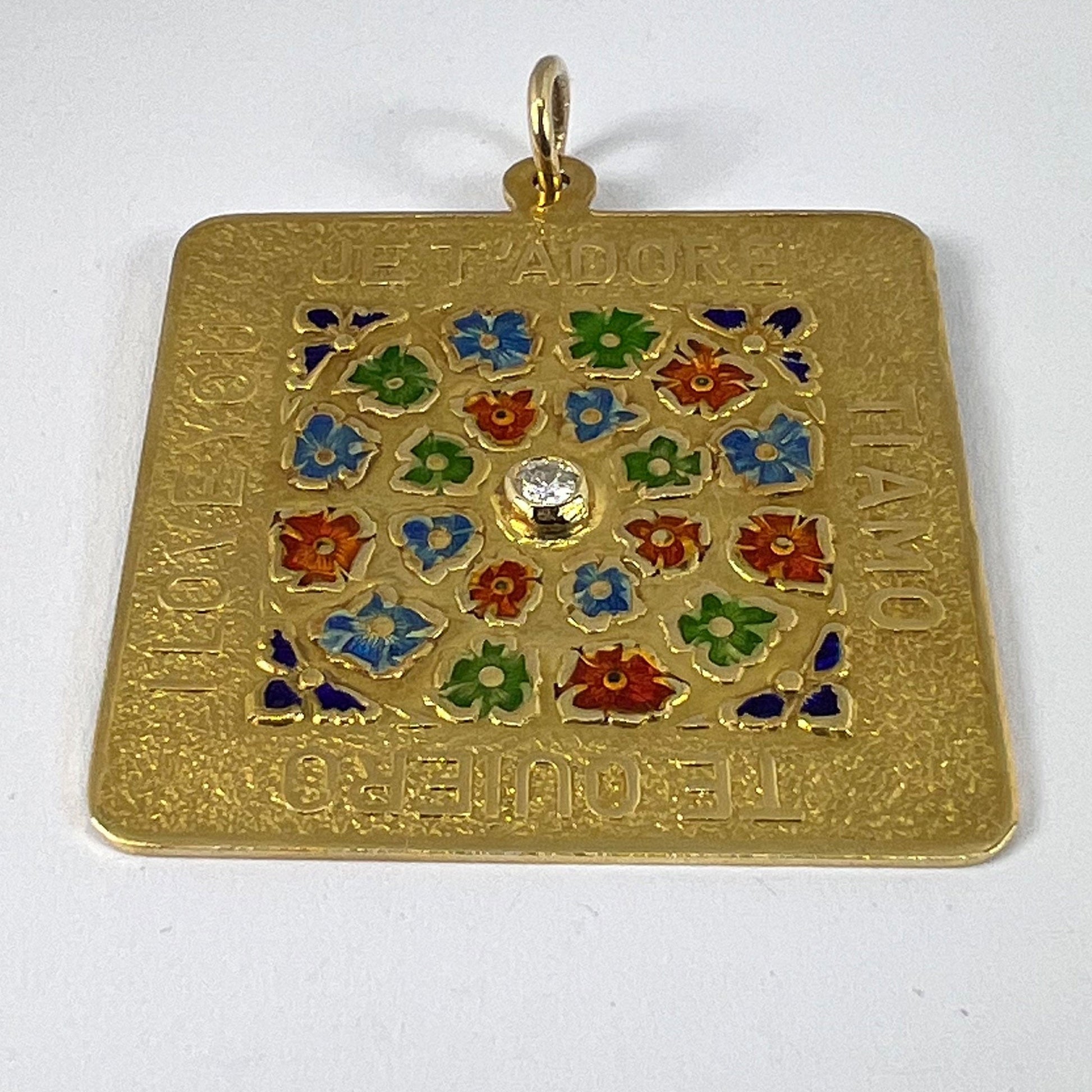 A square shaped yellow gold pendant with enamelled flowers and a central diamond, with text reading JE T'ADORE, TI AMO, TE QUIERO, I LOVE YOU