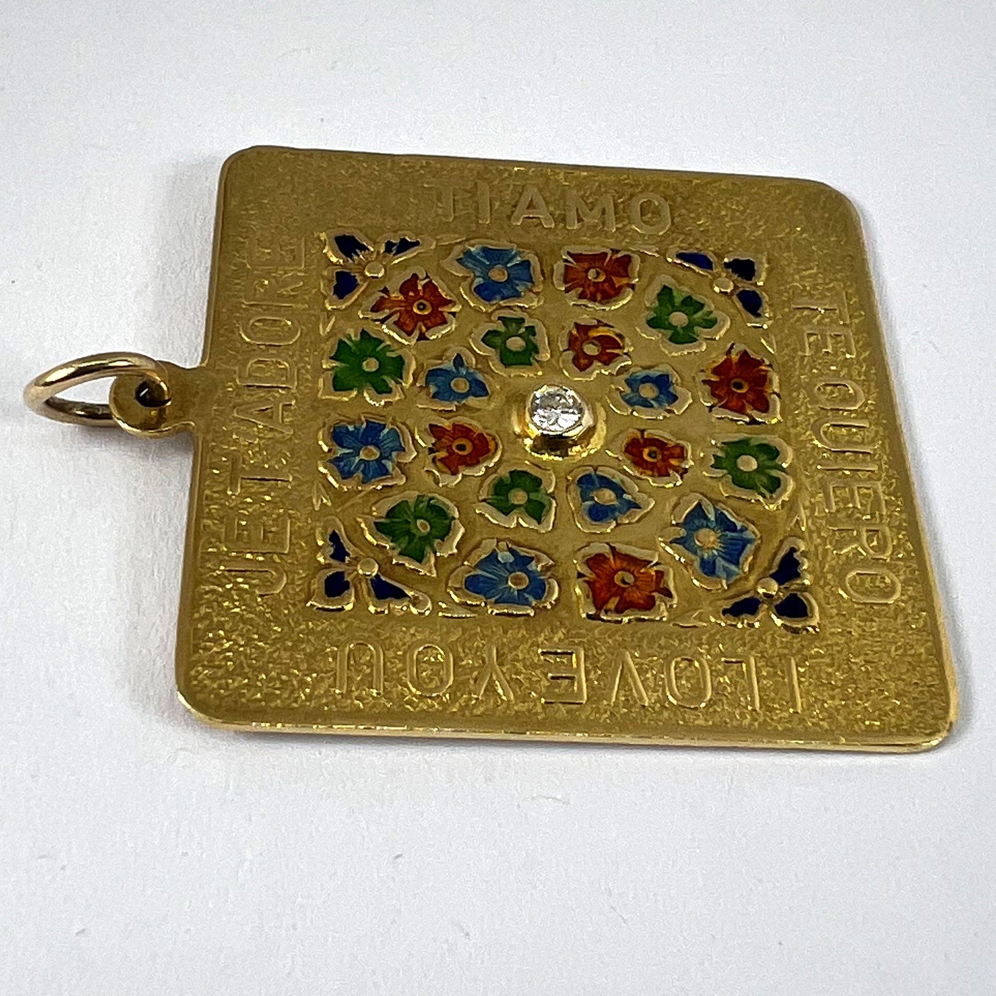 A square shaped yellow gold pendant with enamelled flowers and a central diamond, with text reading JE T'ADORE, TI AMO, TE QUIERO, I LOVE YOU