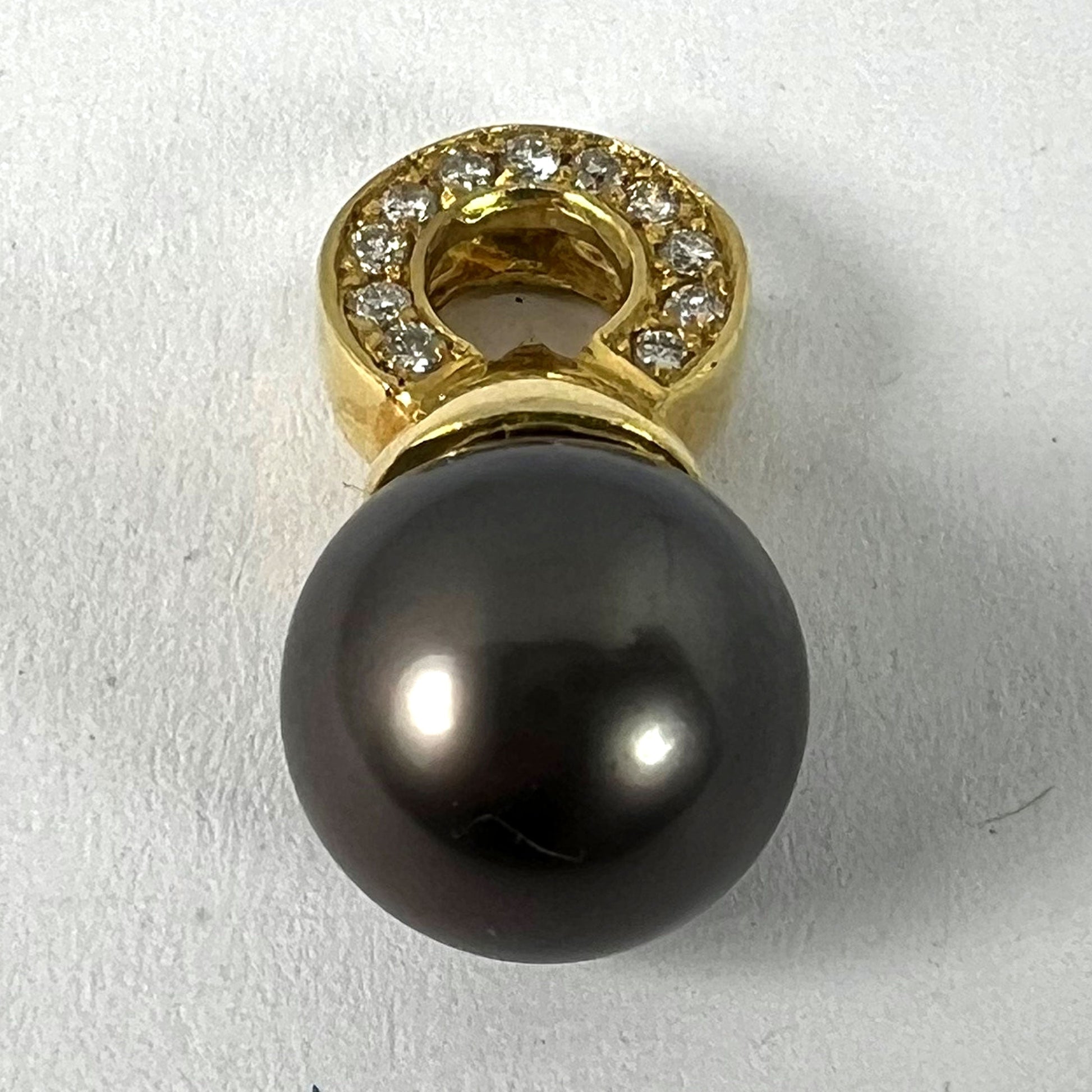 A black pearl pendant with a yellow gold bale set with diamonds, on a white background