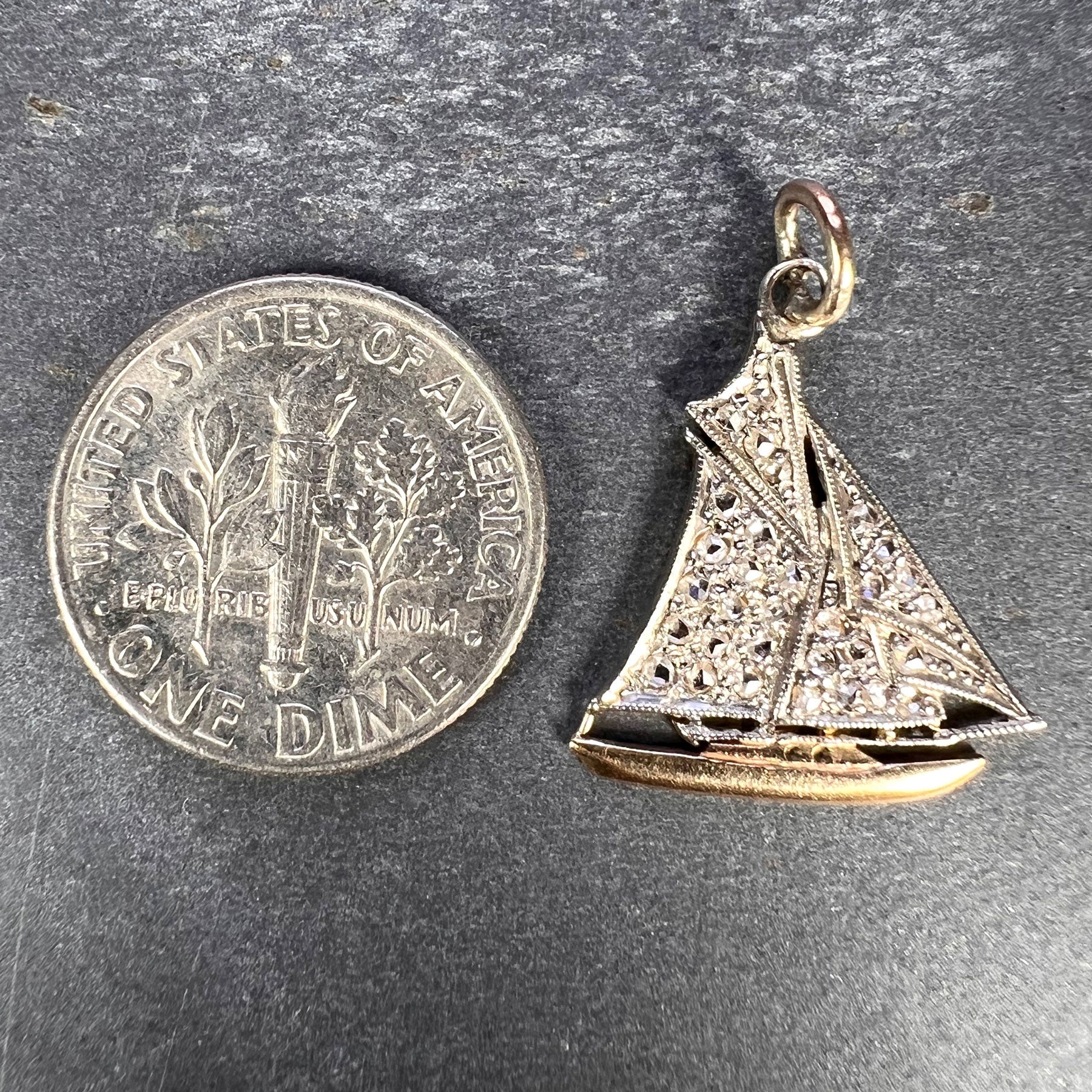 A diamond set yacht boat pendant, next to a dime coin on a grey background