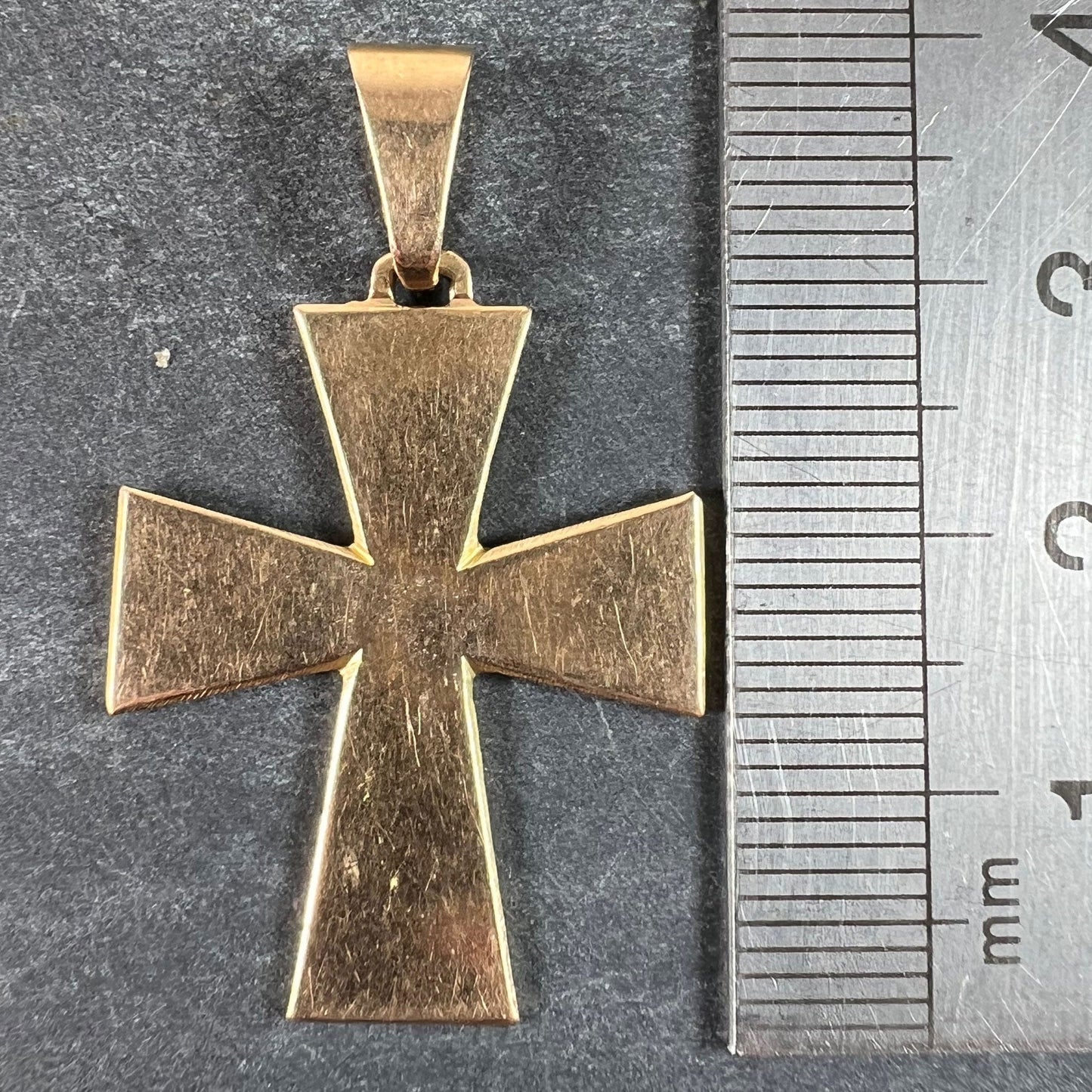 A plain gold cross pendant, next to a metal ruler on a grey background