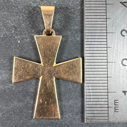A plain gold cross pendant, next to a metal ruler on a grey background