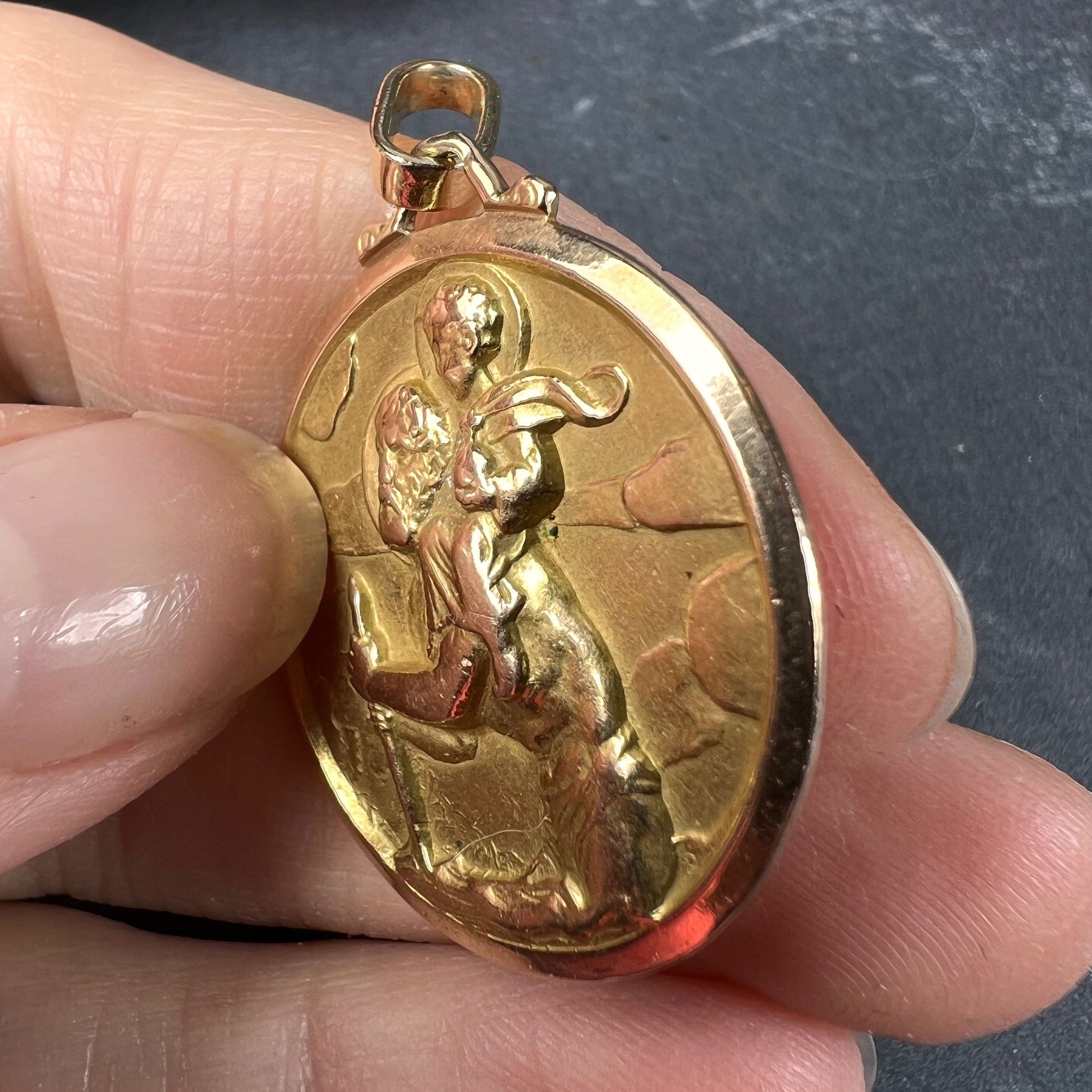 A round yellow gold Saint Christopher pendant, held in a hand on a grey background