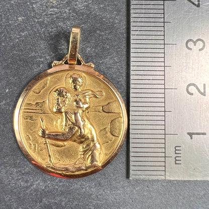 A round yellow gold Saint Christopher pendant, next to a metal ruler on a grey background