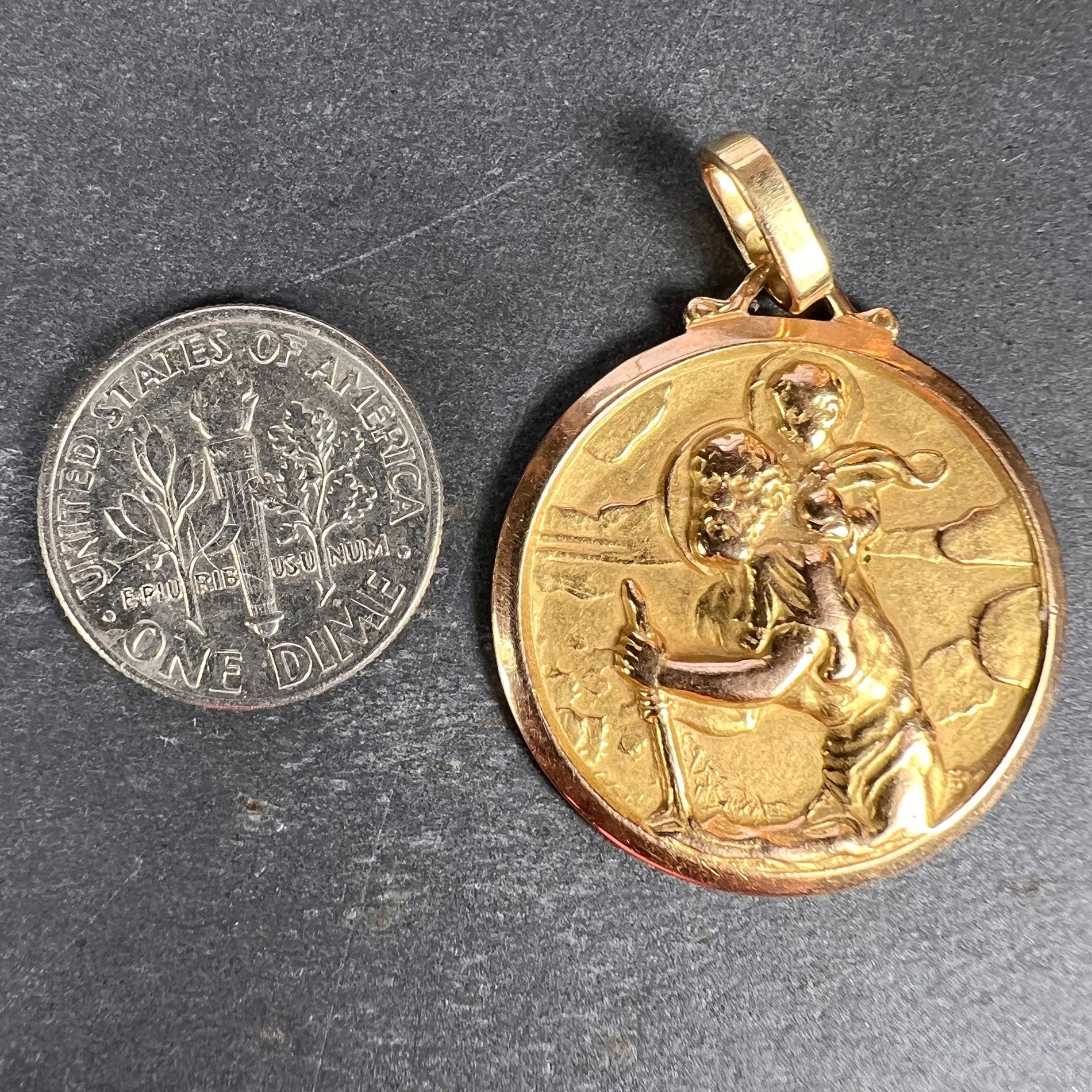A round yellow gold Saint Christopher pendant, next to a dime coin on a grey background
