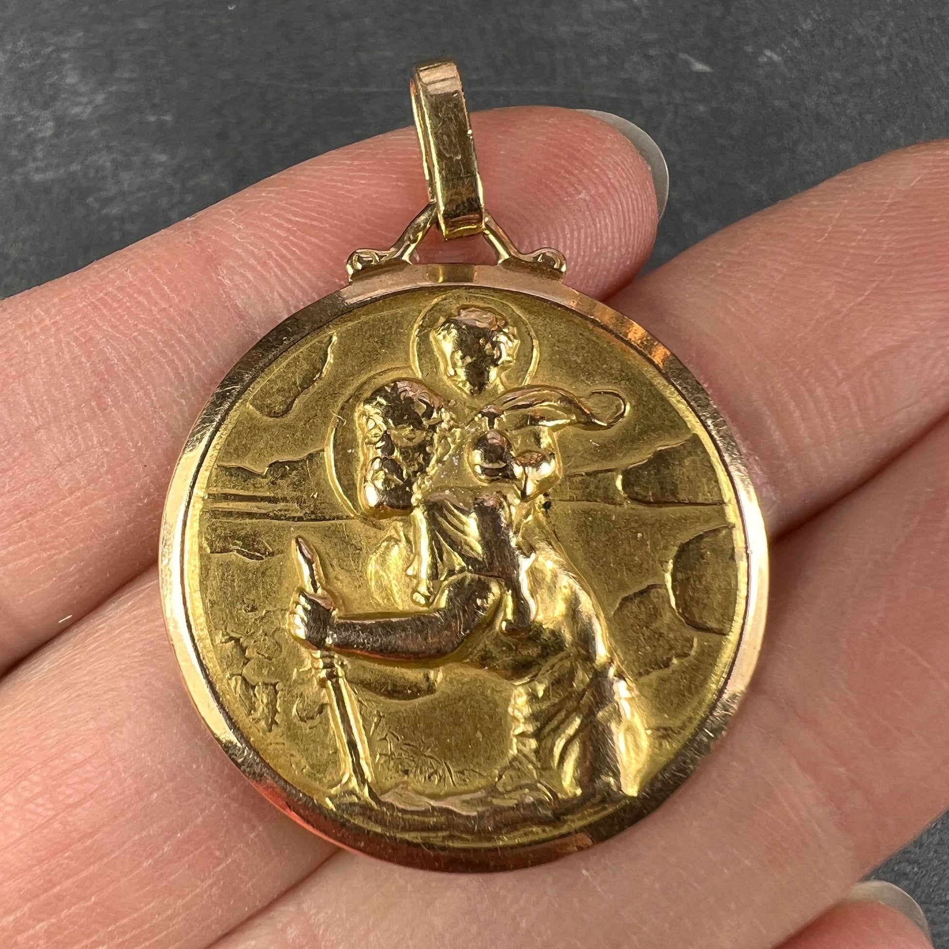 A round yellow gold Saint Christopher pendant, held in a hand on a grey background