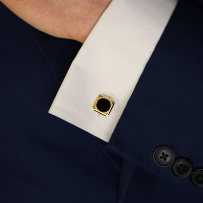 A yellow gold cufflink with circular black onyx on a white cuff, against a dark blue suit